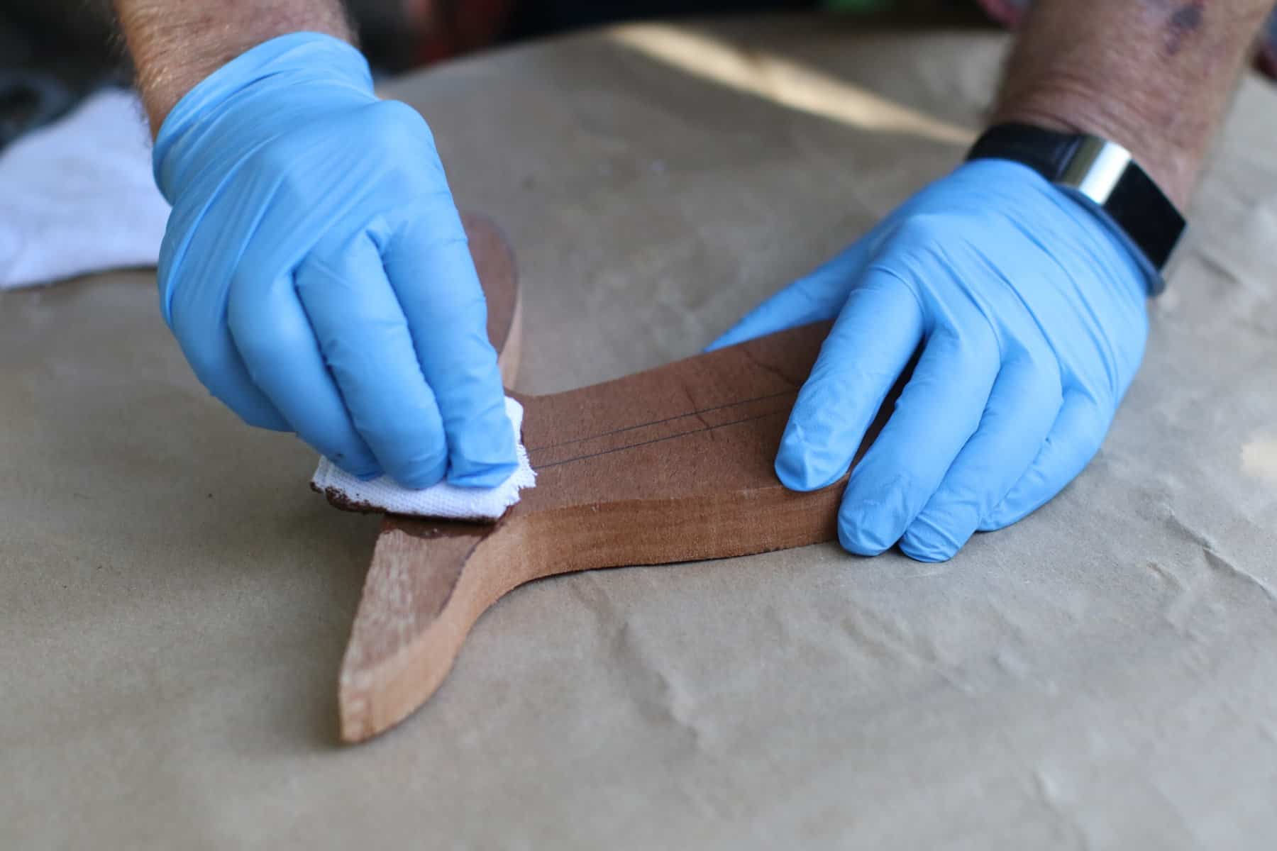 A person wearing blue latex gloves is wiping a brown stain on a piece of wood on top of a brown paper